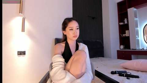 Media: Video of a young Asian woman with long black hair, sitting on a bed in a modern, minimalist room. She wears a black halter top and a white bathrobe, with makeup and earrings.