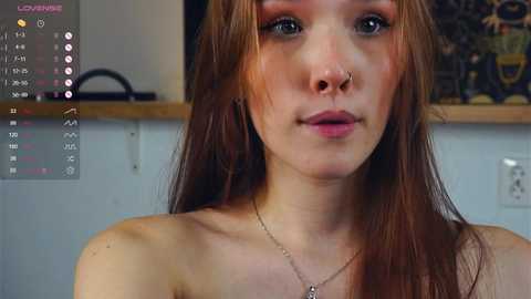 Media: A video of a pale-skinned woman with long, auburn hair, wearing a nose ring and minimal makeup, in a dimly lit room with a digital screen displaying her stats.