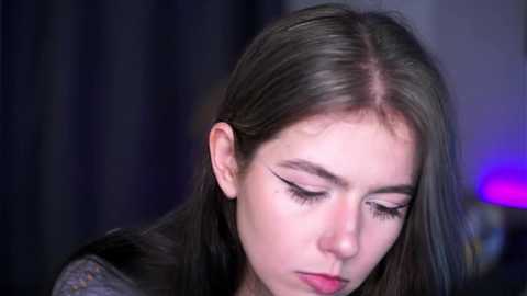 Media: A close-up video of a young Caucasian woman with long, straight, dark brown hair, wearing black eyeliner and mascara, looking down with a serious expression.