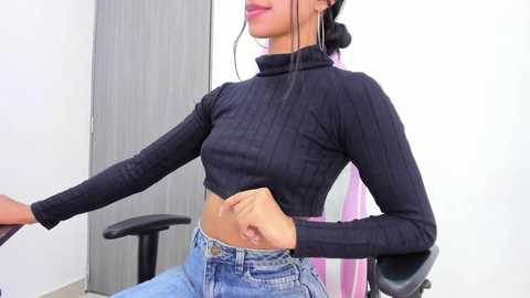 Media: Video of a woman with medium skin tone, wearing a black ribbed crop top and high-waisted jeans, sitting on a black office chair in a minimalist room with gray walls.