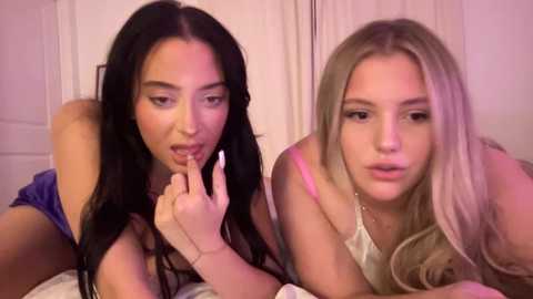 Media: Video of two young women with long hair, one with dark hair and the other with blonde, both wearing lingerie, lying on a bed in a dimly lit room.