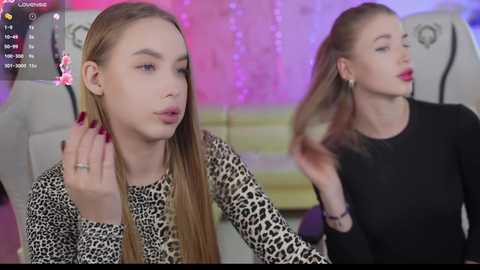 Media: Video of two young women with fair skin and long hair, one with straight, light brown hair and the other with straight, dark brown hair, both wearing black tops, seated in a cozy, dimly lit room with purple and pink fairy lights.