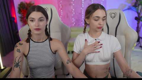 Media: Video of two young women with pale skin, straight brown hair in pigtails, wearing striped and white crop tops, sitting on gaming chairs in a colorful, modern room.