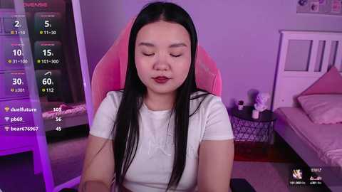 Media: A video of an Asian woman with long black hair, wearing a white shirt, sitting in a pink gaming chair in a dimly lit room.