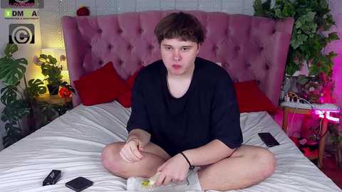 Media: Video of a young, pale-skinned man with short brown hair, wearing a black shirt, sitting cross-legged on a bed with a pink headboard. He's holding a phone and has a pained expression. The room has a green plant, a red pillow, and a laptop on the bed.