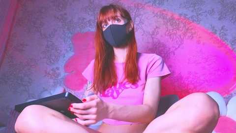 A video of a fair-skinned, red-haired woman in a pink t-shirt and mask, sitting cross-legged on a bed with floral wallpaper, holding a phone.