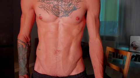 Media: Video of a shirtless, muscular man with fair skin, visible tattoos on chest, arms, and abdomen, standing in a dimly lit room with a wooden chair and a window in the background.