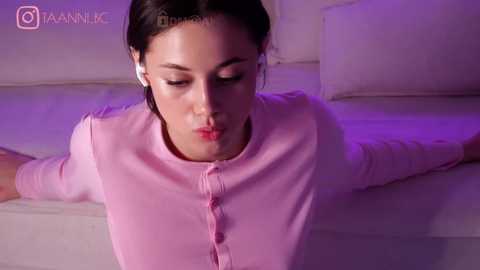 Media: Video of a young woman with olive skin and straight brown hair, wearing pink headphones and a light pink cardigan, lying on a white bed in a dimly lit room.