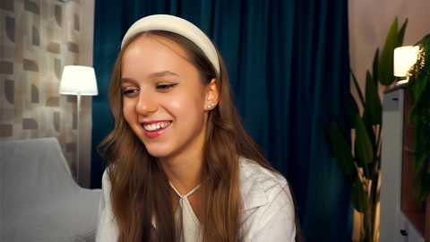 Media: Video of a young girl with light skin, long brown hair, and a white headband, smiling. She wears a beige top, in a modern room with teal curtains, a white chair, and a potted plant.