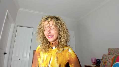 Media: A video of a smiling, curly-haired woman wearing a vibrant yellow and orange dress, standing in a minimalist, brightly lit room with white walls and closed doors.