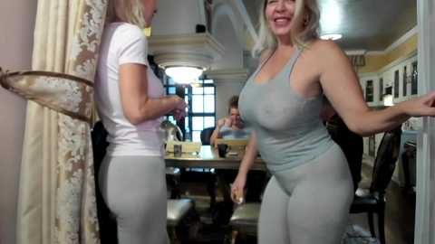 Media: Video of two women, one in a white t-shirt, the other in a light gray tank top and pants, smiling, in a warmly lit, elegant dining room with a chandelier and large windows.