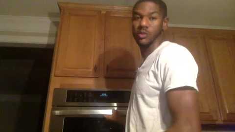 Media: Video of a Black man with short hair, wearing a white T-shirt, standing in a kitchen with wooden cabinets and stainless steel ovens.