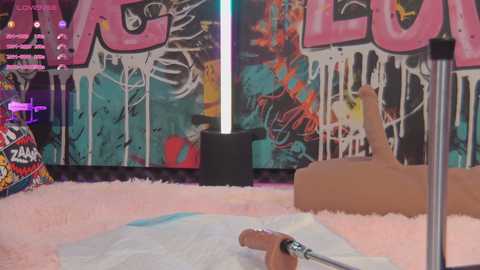 Media: Video of a young woman with a light tan complexion lying on a pink shag rug, holding a sex toy, in a room with vibrant graffiti wall art and a neon light.