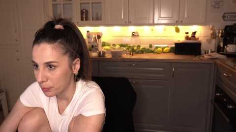 Media: A video of a young woman with a ponytail and white top sitting in a modern kitchen with white cabinets, green vegetables on the counter, and a yellow light background.