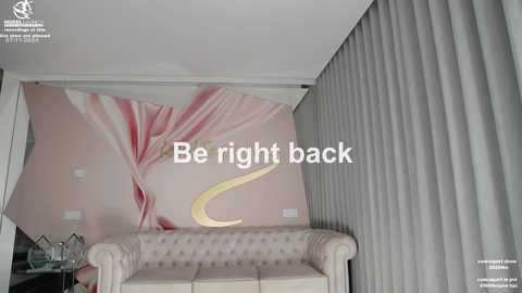 Media: Video of a modern, pink-themed room with a large, abstract floral mural, a tufted beige sofa, and grey vertical curtains. Text overlay reads: \"Be right back.\