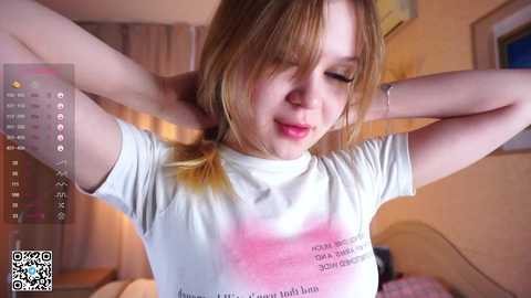 Media: Video of a young Caucasian woman with light brown hair, wearing a white t-shirt, adjusting her hair in a cozy bedroom.