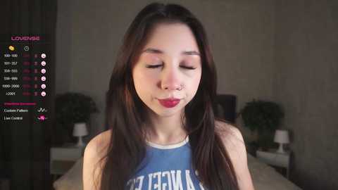 Media: Video of a young Asian woman with long black hair, wearing a blue tank top with white text, and red lipstick, in a dimly lit bedroom with a bed and potted plant in the background.