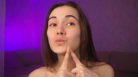 Media: Video of a topless, fair-skinned young woman with straight brown hair, making a peace sign with her fingers against a purple wall and a couch.