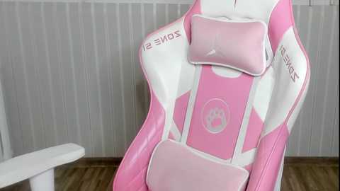 Media: Video of a pink and white gaming chair with a cat paw logo, plush pink headrest, and white arms with \"SOMA\" branding, set against a light grey vertical striped wall and white table.