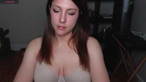 Media: A video of a young woman with fair skin and long brown hair, wearing a beige lace bra, sitting indoors with dim lighting.