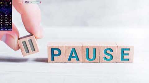 Media: Video of a hand holding a wooden block with \"PAUSE\" in blue letters, placed on a white surface. In the background, a smartphone screen displays code.