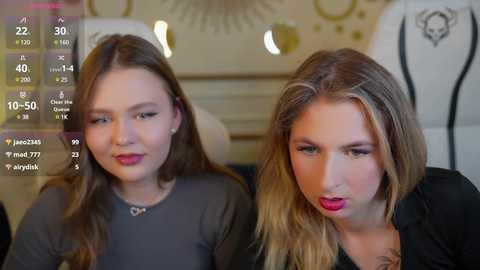 Media: Video of two teenage girls with long hair, wearing makeup, seated indoors. One wears a black top, the other a gray top, both with makeup and lipstick. Background features a mannequin and jewelry display.