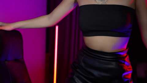 Media: Video of a woman in a black strapless bandeau top and high-waisted leather skirt, standing against a purple and red-lit backdrop.