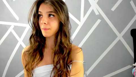 Media: Video of a young woman with long, wavy brown hair, wearing a white off-shoulder top, standing against a geometric, white-patterned gray wall.