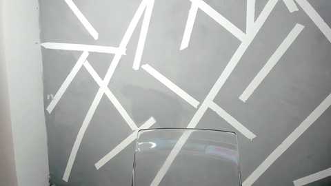 Media: Video of a minimalist interior featuring a grey wall with white, intersecting arrow lines, creating a modern, abstract pattern. A clear glass table sits at the bottom center.