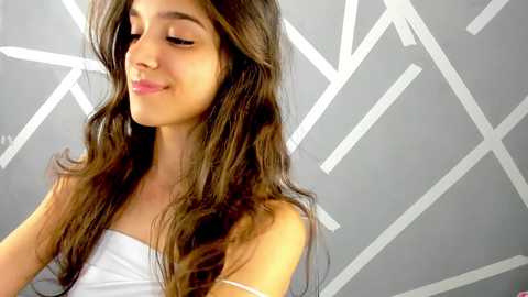 Media: Video of a young woman with long, wavy brown hair, fair skin, and closed eyes, wearing a white strapless top. Background features a modern geometric white pattern on a grey wall.