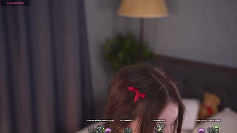 Media: A video of a young woman with brown hair, wearing a red hair bow, lying on a bed with a beige lamp and gray curtains in the background.