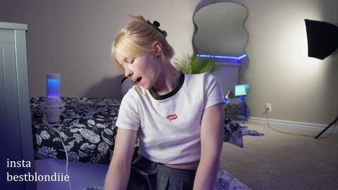 Media: Video of a young blonde woman in a white crop top, sitting on a bed with a black and white floral pattern, in a dimly lit, modern bedroom.