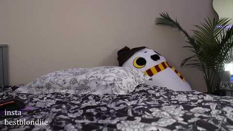 Media: Video of a plush owl with large yellow eyes, a brown hat, and a yellow and red scarf, placed on a bed with a black and white floral duvet, against a beige wall.