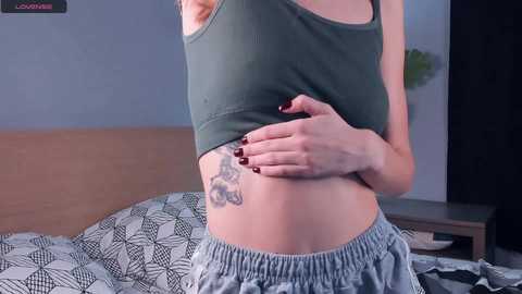 Media: A video of a slender, fair-skinned woman with small breasts, wearing a green crop top and loose gray shorts. She has a floral tattoo on her right hip, standing in a bedroom with a patterned bedspread.