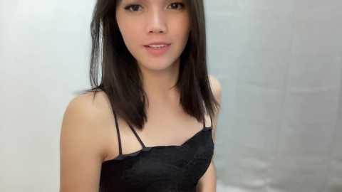 Media: Video of a young Asian woman with straight, dark brown hair and light skin, wearing a black lace top with thin straps. She has a neutral expression and is standing against a blurred, light-colored background.