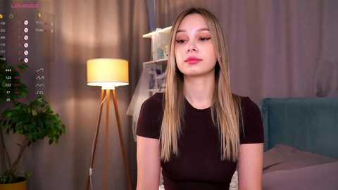 Media: Video of a young woman with straight blonde hair, fair skin, and a slim figure, wearing a black short-sleeve top. Background includes a lamp, a teal couch, and a potted plant.