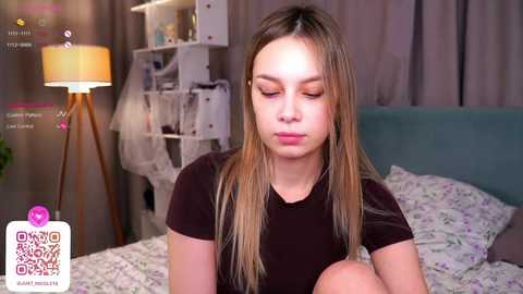 Media: Video of a young woman with long blonde hair, wearing a black t-shirt, sitting on a bed in a cozy bedroom with a white lamp, floral bedding, and a shelf in the background.