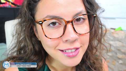 Media: Video of a smiling, light-skinned woman with curly brown hair and large, brown-framed glasses, wearing a green top, sitting indoors on a stone floor.