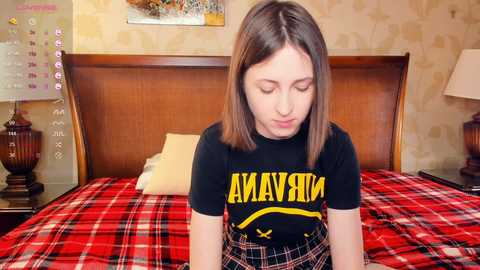 Video of a young Caucasian woman with straight brown hair, wearing a black t-shirt with yellow text and plaid skirt, sitting on a bed with red and black checkered sheets in a vintage-style bedroom.