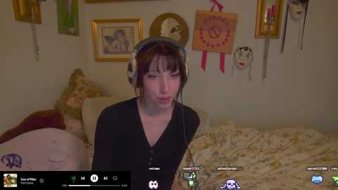 Media: A video of a pale-skinned woman with dark hair in a black top, wearing headphones, in a cluttered, dimly lit bedroom with framed pictures and a bed.