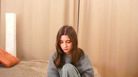 Media: Video of a young woman with straight brown hair, wearing a gray sweater and light jeans, sitting on a textured beige couch, with beige curtains in the background.