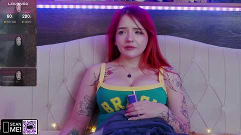 Media: Video of a tattooed, red-haired woman in a green tank top with \"Brasil\" written on it, sitting on a cream-colored, tufted couch, holding a cigarette, in a dimly lit room.