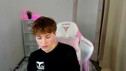 Media: Video of a young man with short brown hair, wearing a black t-shirt with a white logo, seated in a pink and white gaming chair, surrounded by a simple, brightly lit room with white walls and a small, purple-lit plant on a shelf.