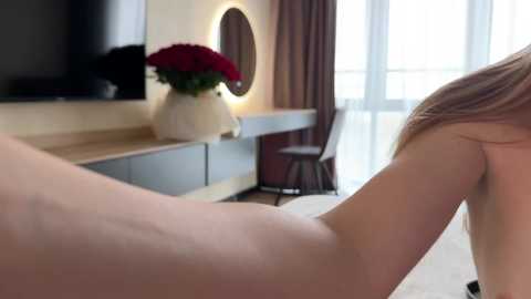 Media: Video of a naked woman with fair skin, lying face-down on a bed, her left arm raised, in a modern bedroom with a white desk, vase of red roses, and large window.