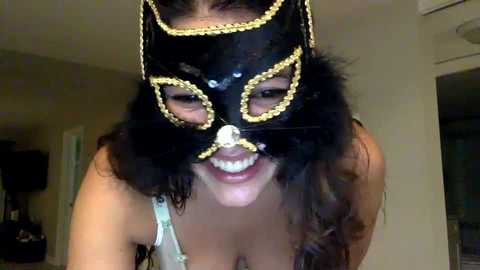 Media: Video of a smiling woman in a black, sequined masquerade mask with yellow beading, wearing a white lace bra, indoors with beige walls and a door visible in the background.