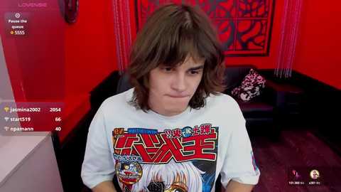 Media: Video of a young man with shoulder-length brown hair, wearing a white T-shirt with a colorful anime character design, in a red-lit room with black furniture and a Japanese-style screen.