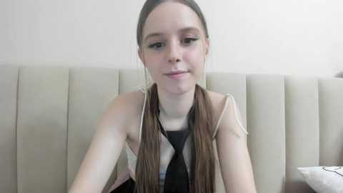 Media: Video of a young Caucasian girl with long brown pigtails, wearing a black and white striped shirt, sitting on a beige upholstered bench against a plain white wall.