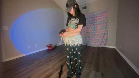 Media: Video of a young woman in a dimly lit room with a rainbow light effect, wearing a black graphic tee, floral pajama pants, and a hat, holding a small stuffed animal.