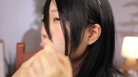 Media: A video of a young Asian woman with long black hair, pink lips, and a light skin tone, licking a man's erect penis in a blurred, intimate indoor setting.