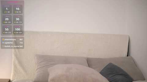 Media: A video of a minimalist bedroom with a beige upholstered headboard, two pillows, and a single gray pillow. The background is a plain white wall with a digital temperature display overlay on the left.